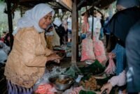 Seorang wanita modern menikmati segelas jamu kunyit asam segar di sebuah kafe jamu kekinian, menunjukkan bagaimana minuman tradisional ini telah beradaptasi dengan gaya hidup urban. Berbagai rempah seperti jahe, kunyit, dan kencur yang menjadi bahan dasar jamu tampak tertata rapi di latar belakang, mengisyaratkan kekayaan alam Indonesia.
