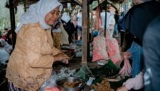 Seorang wanita modern menikmati segelas jamu kunyit asam segar di sebuah kafe jamu kekinian, menunjukkan bagaimana minuman tradisional ini telah beradaptasi dengan gaya hidup urban. Berbagai rempah seperti jahe, kunyit, dan kencur yang menjadi bahan dasar jamu tampak tertata rapi di latar belakang, mengisyaratkan kekayaan alam Indonesia.