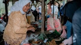 Seorang wanita modern menikmati segelas jamu kunyit asam segar di sebuah kafe jamu kekinian, menunjukkan bagaimana minuman tradisional ini telah beradaptasi dengan gaya hidup urban. Berbagai rempah seperti jahe, kunyit, dan kencur yang menjadi bahan dasar jamu tampak tertata rapi di latar belakang, mengisyaratkan kekayaan alam Indonesia.