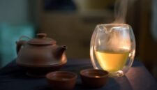Secangkir teh hijau hangat mengepulkan uap, siap dinikmati untuk memulai hari dengan penuh energi dan manfaat kesehatan. Daun teh hijau segar terlihat di latar belakang, melambangkan kealamian dan kekayaan nutrisi minuman ini.