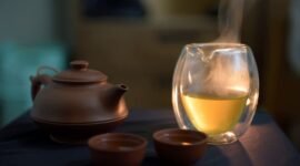 Secangkir teh hijau hangat mengepulkan uap, siap dinikmati untuk memulai hari dengan penuh energi dan manfaat kesehatan. Daun teh hijau segar terlihat di latar belakang, melambangkan kealamian dan kekayaan nutrisi minuman ini.