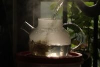 Secangkir teh hijau hangat mengepul di pagi hari, dikelilingi daun teh segar, menggambarkan kehangatan dan kesegaran yang ditawarkan minuman kaya manfaat ini.