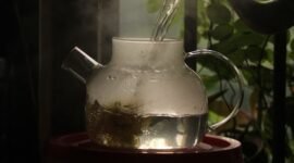 Secangkir teh hijau hangat mengepul di pagi hari, dikelilingi daun teh segar, menggambarkan kehangatan dan kesegaran yang ditawarkan minuman kaya manfaat ini.