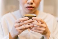 Seorang wanita menikmati segelas jamu kunyit asam yang menyegarkan, menunjukkan perpaduan antara tradisi dan gaya hidup sehat masa kini. Berbagai rempah dan bahan alami, seperti kunyit dan asam jawa, terlihat di sekelilingnya, menggambarkan kekayaan warisan herbal Indonesia.