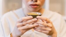 Seorang wanita menikmati segelas jamu kunyit asam yang menyegarkan, menunjukkan perpaduan antara tradisi dan gaya hidup sehat masa kini. Berbagai rempah dan bahan alami, seperti kunyit dan asam jawa, terlihat di sekelilingnya, menggambarkan kekayaan warisan herbal Indonesia.