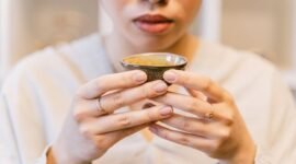 Seorang wanita menikmati segelas jamu kunyit asam yang menyegarkan, menunjukkan perpaduan antara tradisi dan gaya hidup sehat masa kini. Berbagai rempah dan bahan alami, seperti kunyit dan asam jawa, terlihat di sekelilingnya, menggambarkan kekayaan warisan herbal Indonesia.