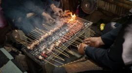 Seorang penjual sedang membuat surabi di atas wajan tanah liat yang dipanaskan menggunakan bara arang, metode tradisional yang memberikan aroma dan cita rasa khas pada kue legendaris ini. Asap tipis yang mengepul menambah nuansa otentik dalam proses pembuatan surabi.